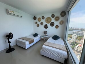 A hotel room with two beds, a small table, a lamp, a fan, and a view of the city.