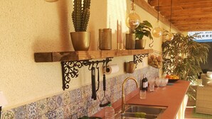 Een keuken met een lange aanrecht, een spoelbak en diverse kookgerei. Er staan potplanten en een hanglamp boven het aanrecht.