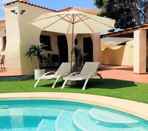 Ein Poolbereich mit zwei Liegestühlen und einem Sonnenschirm, umgeben von einer Terrasse mit Stühlen und Kübelpflanzen.