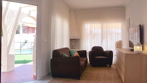 Een woonkamer met een bank, fauteuil en een televisie op een houten dressoir. Er is een raam met gordijnen en een deur naar buiten.