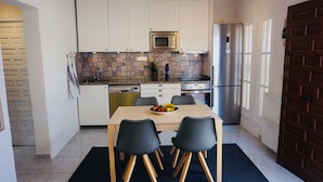Een compacte keuken met witte kastjes, een magnetron, een fornuis en een koelkast. Er staat een eettafel met vier stoelen bij en een schaal fruit erop.