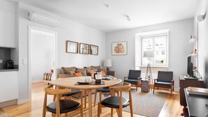 A modern living room with a wooden dining table, black chairs, a sofa, and a coffee table. There is a window with a view of buildings, a lamp, and framed artwork on the wall. - By the Castle | TRENDY COMFY & NEW | Family-Friendly (Lisboa)