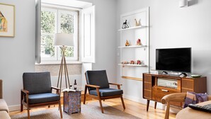 A living room with two armchairs, a wooden coffee table, a TV on a wooden cabinet, and a window with a view of greenery. - By the Castle | TRENDY COMFY & NEW | Family-Friendly (Lisboa)
