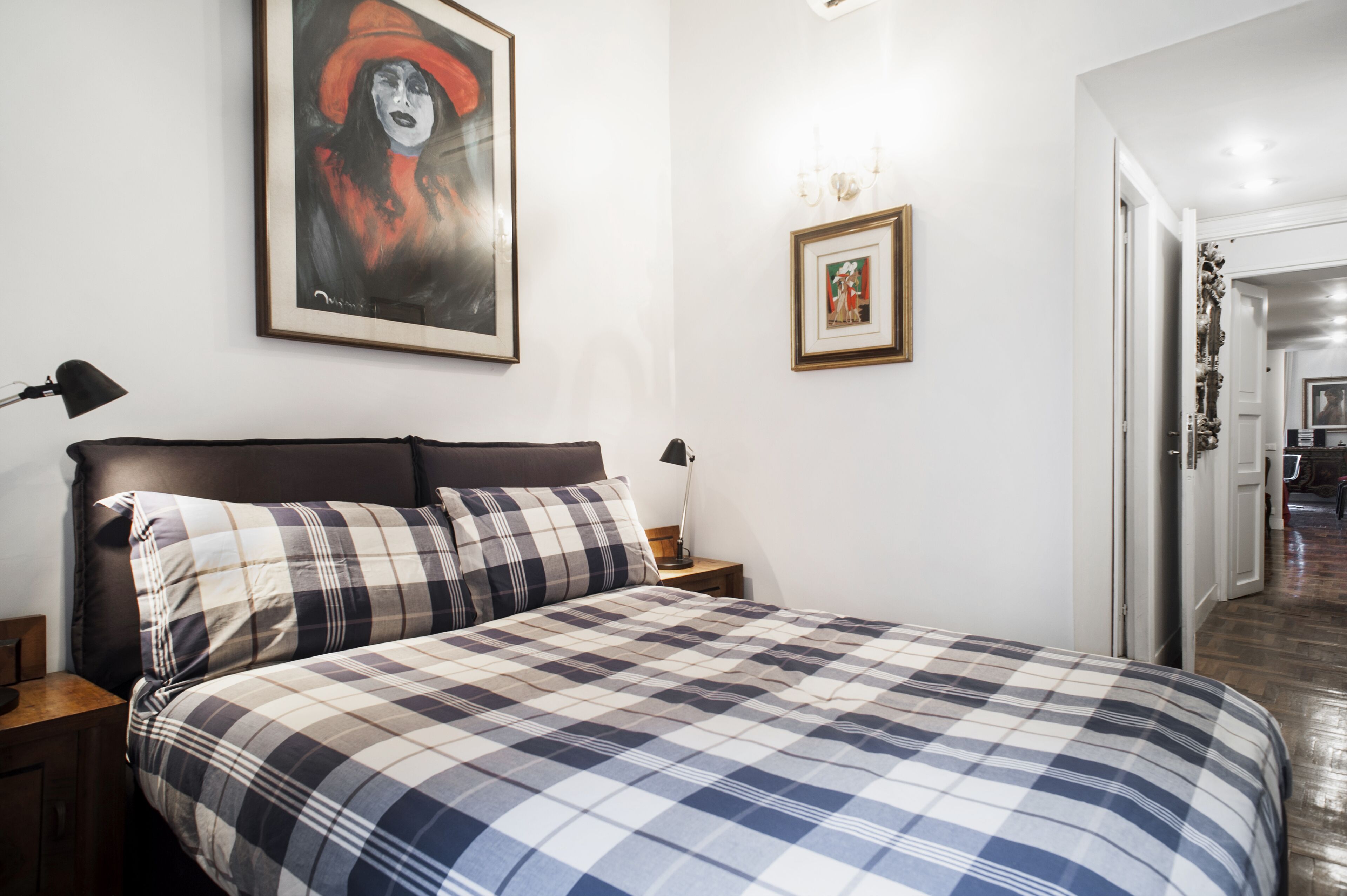 A bedroom with a bed, two bedside lamps, a framed painting, and a framed picture on the wall.
