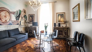 A room with a sofa, a glass table with chairs, a chandelier, and artwork on the walls.