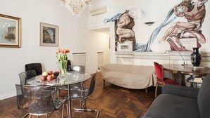 A dining area with a round glass table, clear chairs, a chandelier, and a mural on the wall.