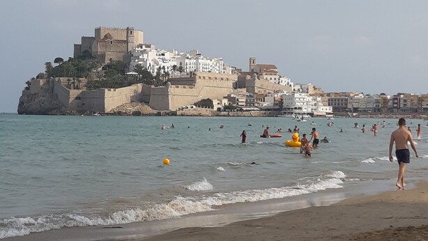 A beach with people swimming and walking, a castle-like structure in the background, and a clear sky.