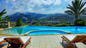 Une piscine à bordures courbes, entourée de chaises longues et de palmiers, avec un paysage montagneux en arrière-plan.