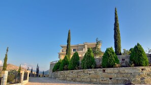 Uma villa com muro de pedra, entrada com calçada e árvores altas.