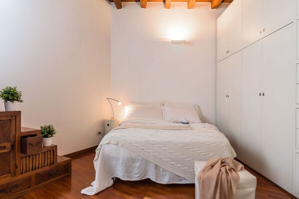 A bedroom with a bed, bedside table, lamp, wooden cabinet, and a potted plant.