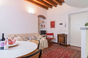 A living room with a sofa, a dining table with chairs, a red patterned rug, a bookshelf, and a television.