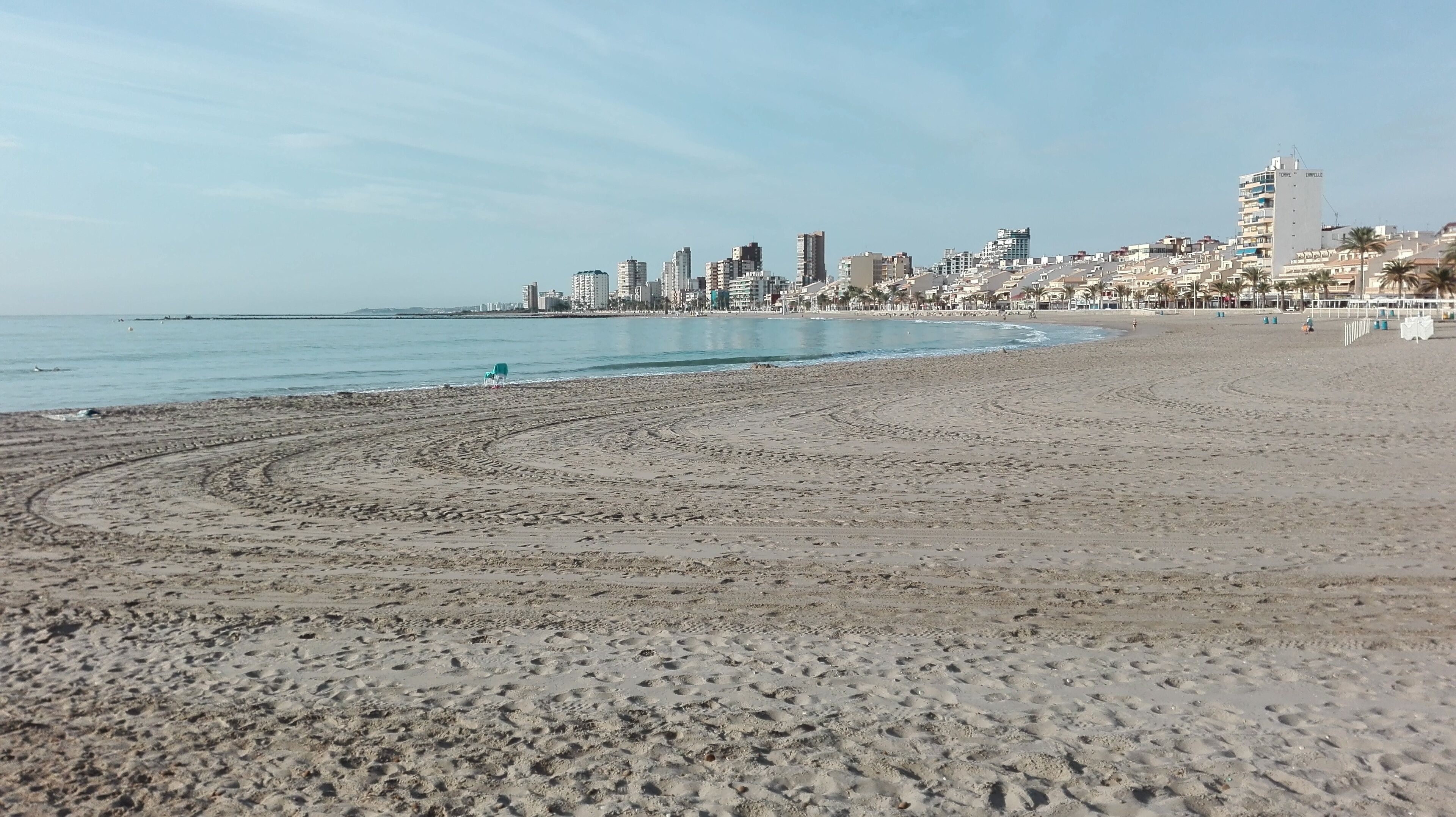 A wide sandy beach with footprints leading to the water, a clear sky, and a city skyline in the background.