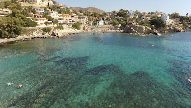 A coastal town with clear turquoise waters, buildings on a hillside, and rocky terrain.