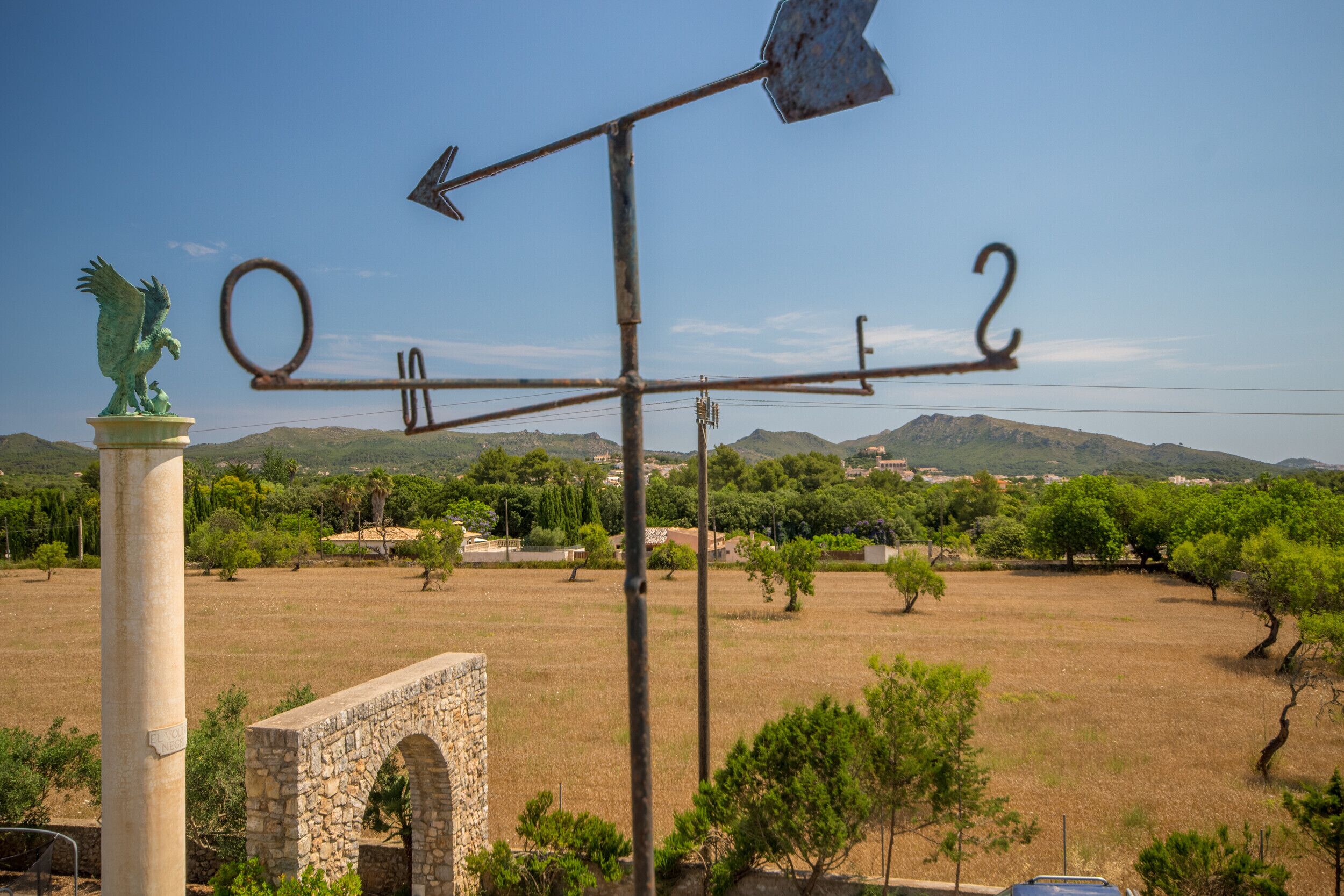 A weather vane with directional arrows and numbers, a stone statue, and an archway in a rural landscape.