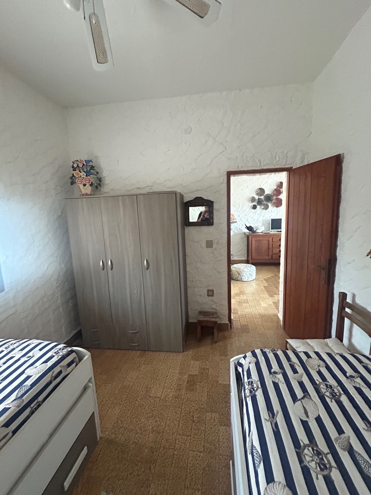A bedroom with two beds, a wardrobe, a small stool, a mirror, and a door leading to another room.