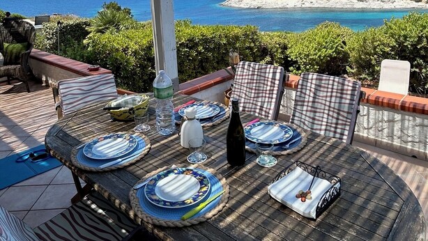 An outdoor dining area with a view of the sea, a pergola, and a table set for two with plates, glasses, and a bottle. - Terrace with dreamy sunsets over the sea! ( CIN code IT111071C2000T8749) (Sant’Antioco)