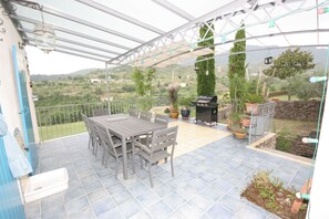 Eine Terrasse im Freien mit Tisch und Stühlen, einem Grill, Blumentöpfen und Blick auf die Landschaft.