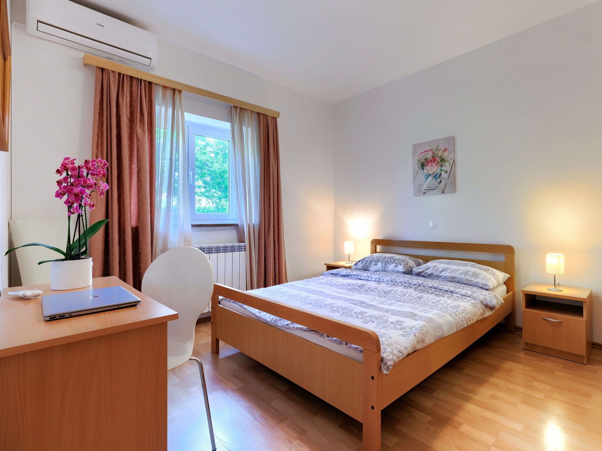 Un dormitorio con una cama de madera, un escritorio con una computadora portátil, una silla, un radiador, una ventana con cortinas y una lámpara de noche.