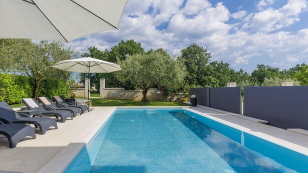 Une piscine entourée de verdure et d’un mur de pierre, avec des chaises longues et un parasol.
