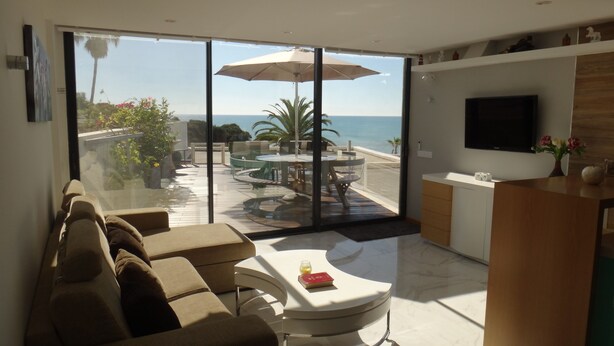 Una sala de estar moderna con un amplio ventanal corredero que da a un patio exterior con piscina, zona de comedor y un televisor montado en la pared.