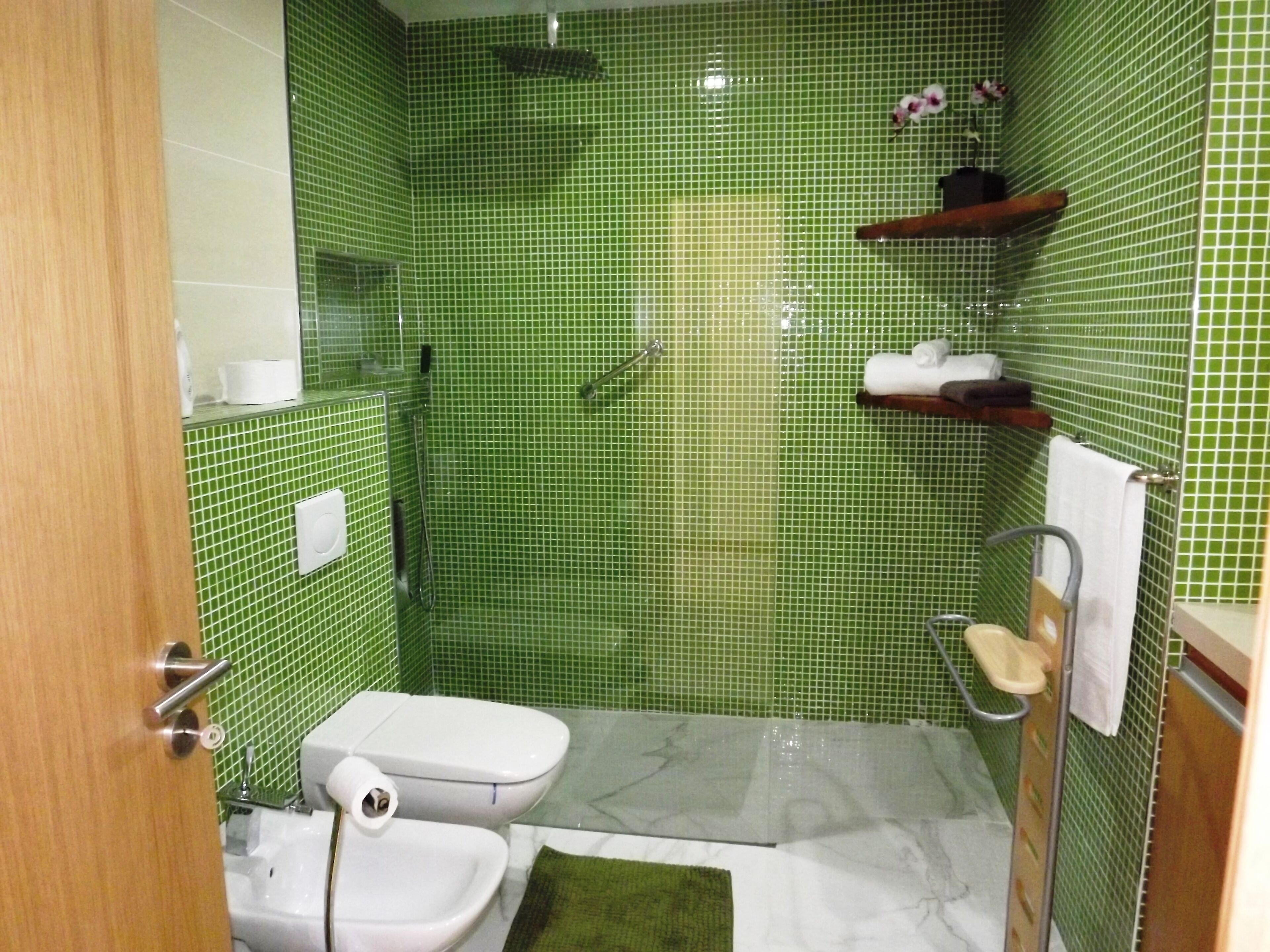A bathroom with green tiled walls, a white toilet, a shower, and a small shelf with towels.