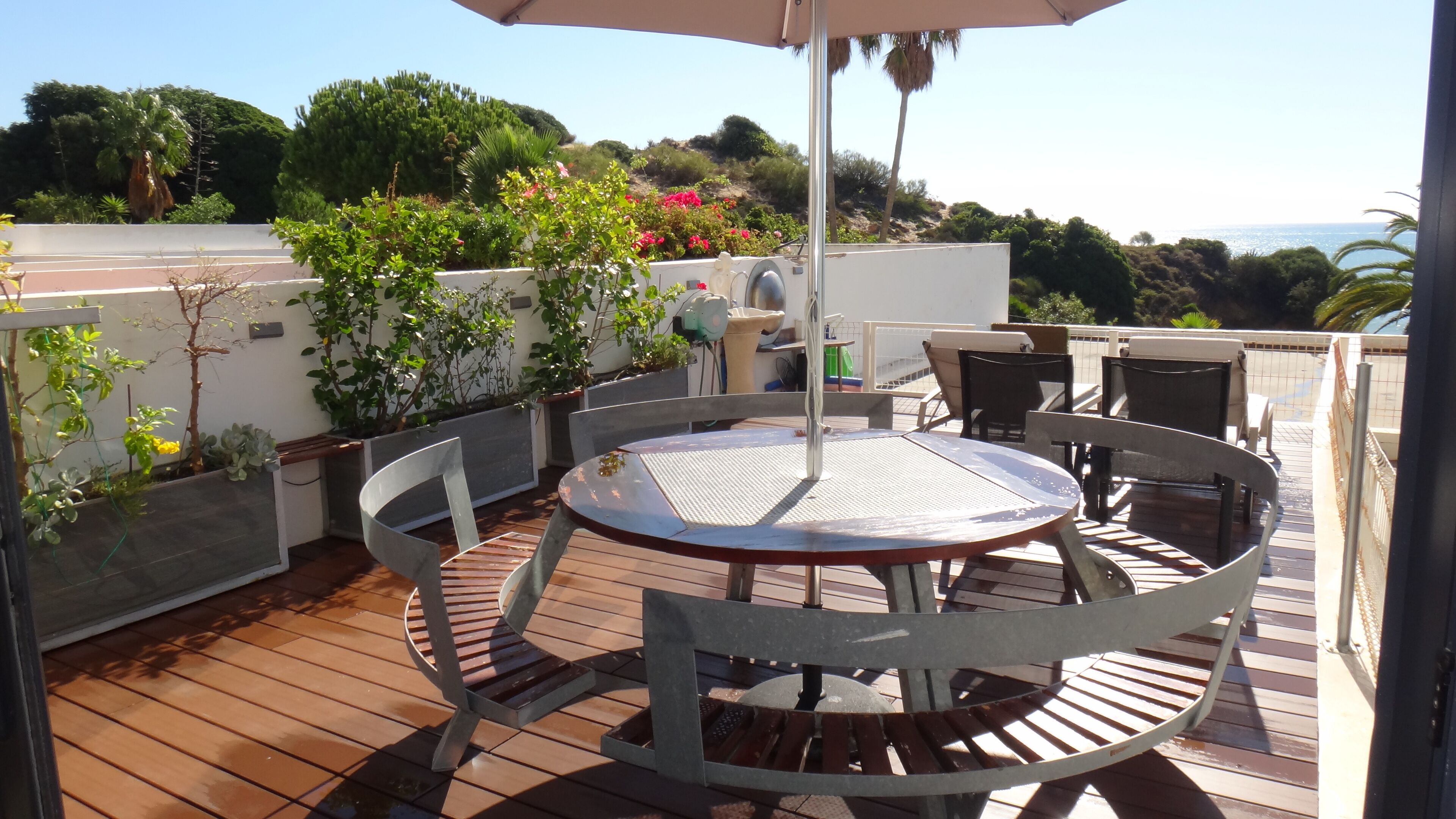 A wooden deck with a round table and chairs, surrounded by potted plants and a view of the sea.