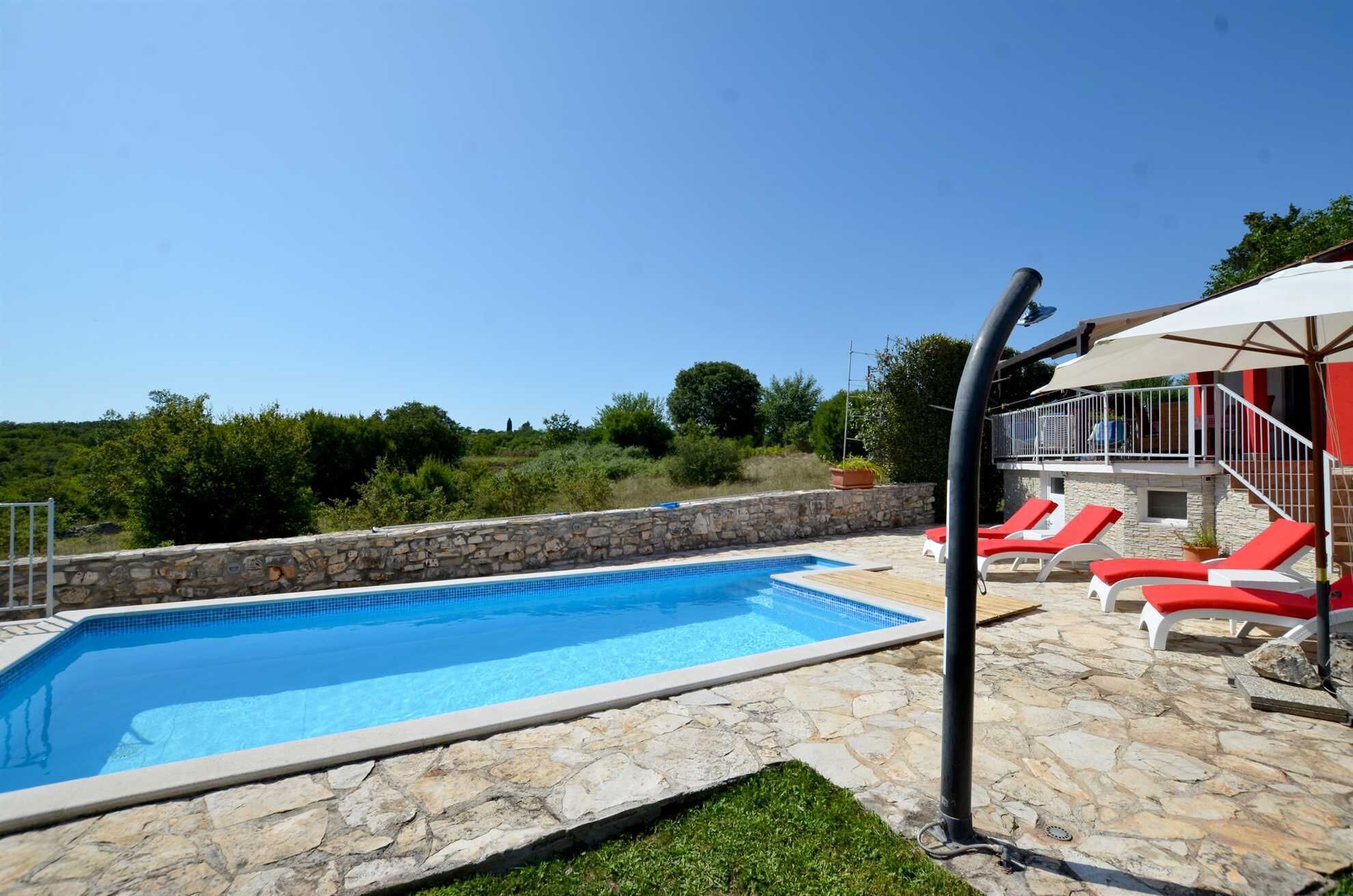 Piscina con baldosas de piedra, sillas reclinables rojas y una pared de piedra.