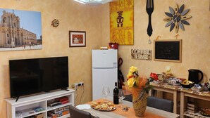Ein Wohnzimmer mit Fernseher, Esstisch, Stühlen, Deckenventilator, Kunstwerken und einem Kühlschrank.