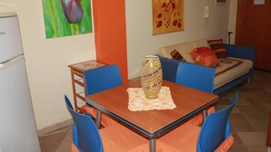 Ein Essbereich mit Tisch, Stühlen und einer dekorativen Vase. Außerdem gibt es einen Kühlschrank, eine Couch und eine Wand mit Wandteppich mit Blumenmuster.