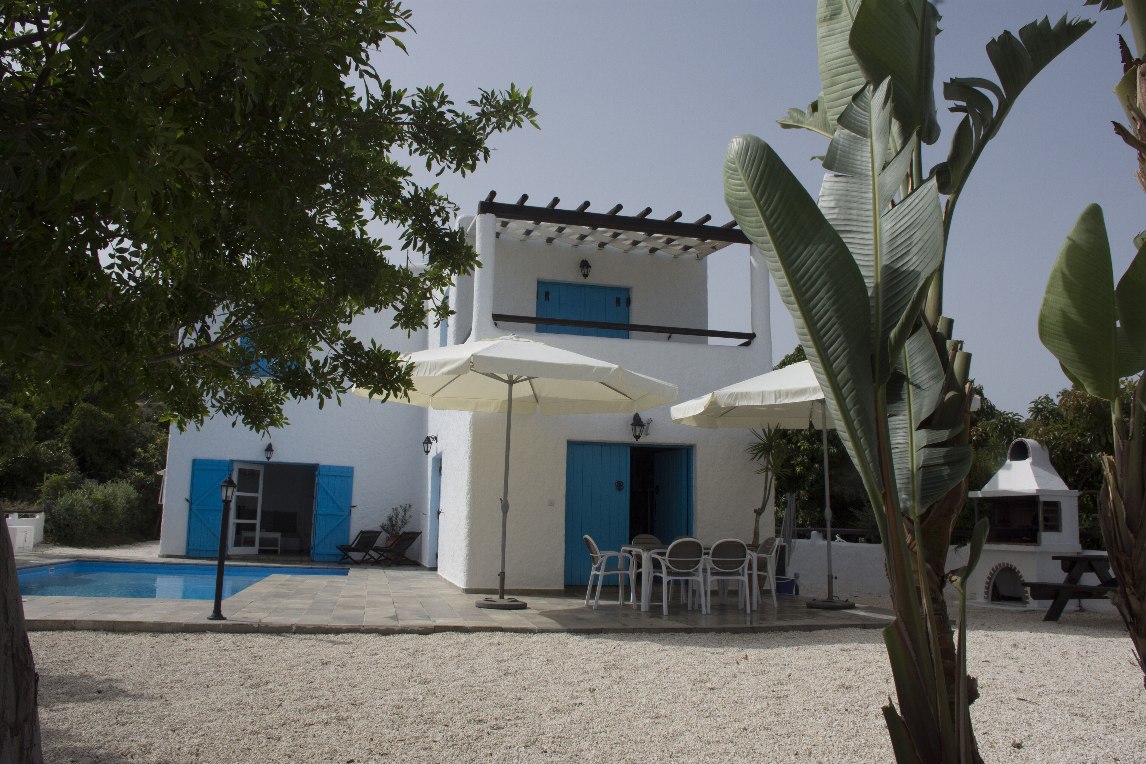 A white building with blue doors and windows, a pool, outdoor seating, and a patio area.
