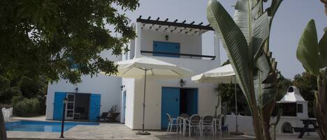 A white building with blue doors and windows, a pool, outdoor seating, and a patio area.