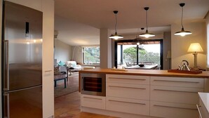 A modern kitchen with a built-in oven, a large island with a sink, pendant lights, and a view of the outdoor patio area. - Modern Villa (Center Of Begur) Overlooking Both Village And Sea (Begur)