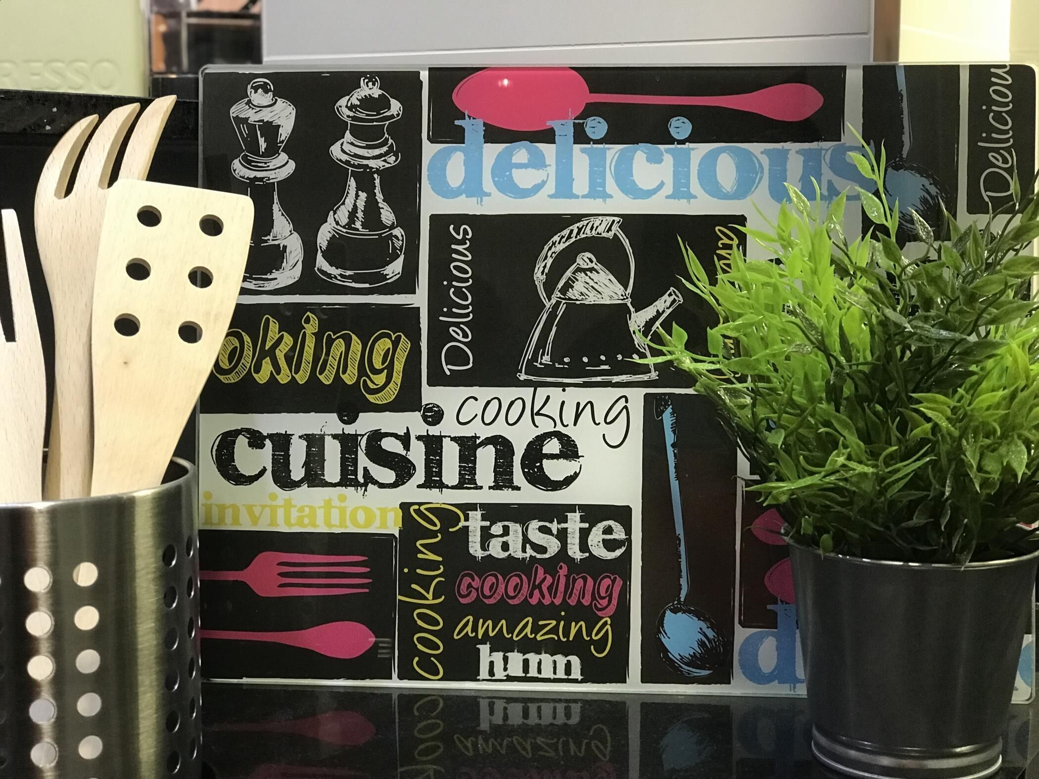 Una encimera de cocina con una pizarra que muestra palabras e ilustraciones relacionadas con la cocina, un recipiente metálico para utensilios y una planta en maceta.