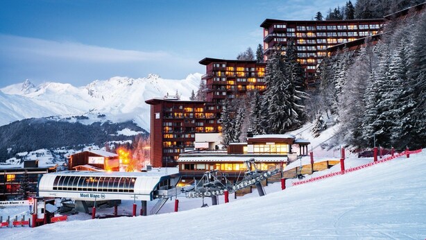 A ski resort with a cable car system, snow-covered mountains, and a building with multiple balconies.
