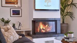 Un salon moderne avec une cheminée, un canapé, une table basse et un téléviseur fixé au-dessus de la cheminée, qui affiche un logo.