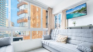 A modern living room with a grey sofa, a flat-screen TV, and a large window offering a view of a high-rise building and the sea. - Inlesa 4-B Apartment Levante Beach (Benidorm)