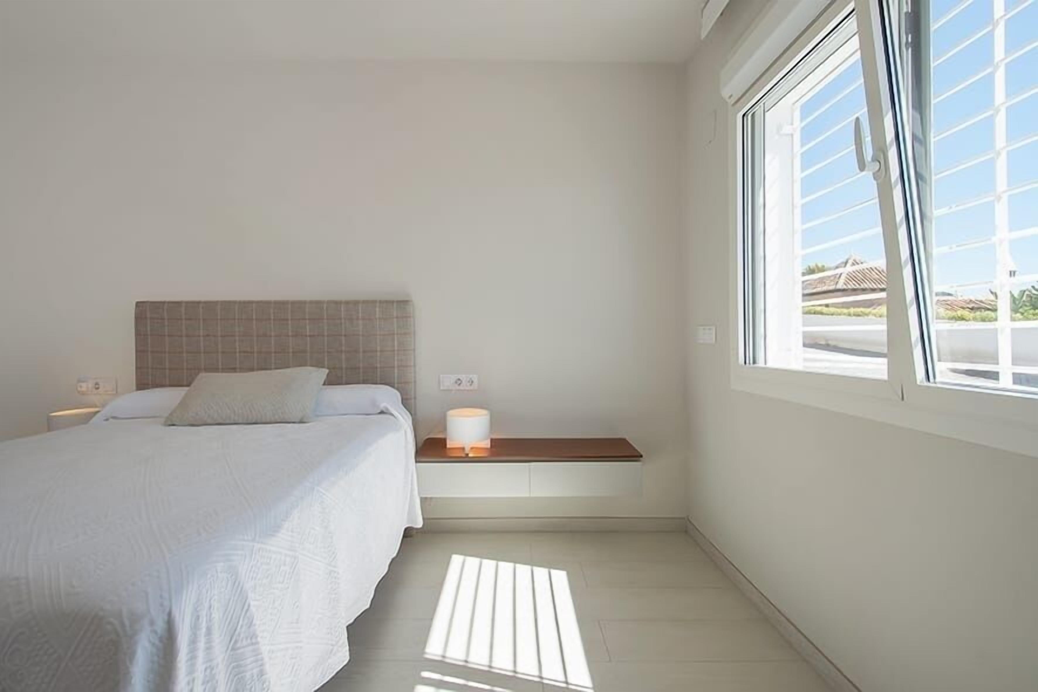Un dormitorio con cama, mesita de noche, una lámpara y vista al exterior.