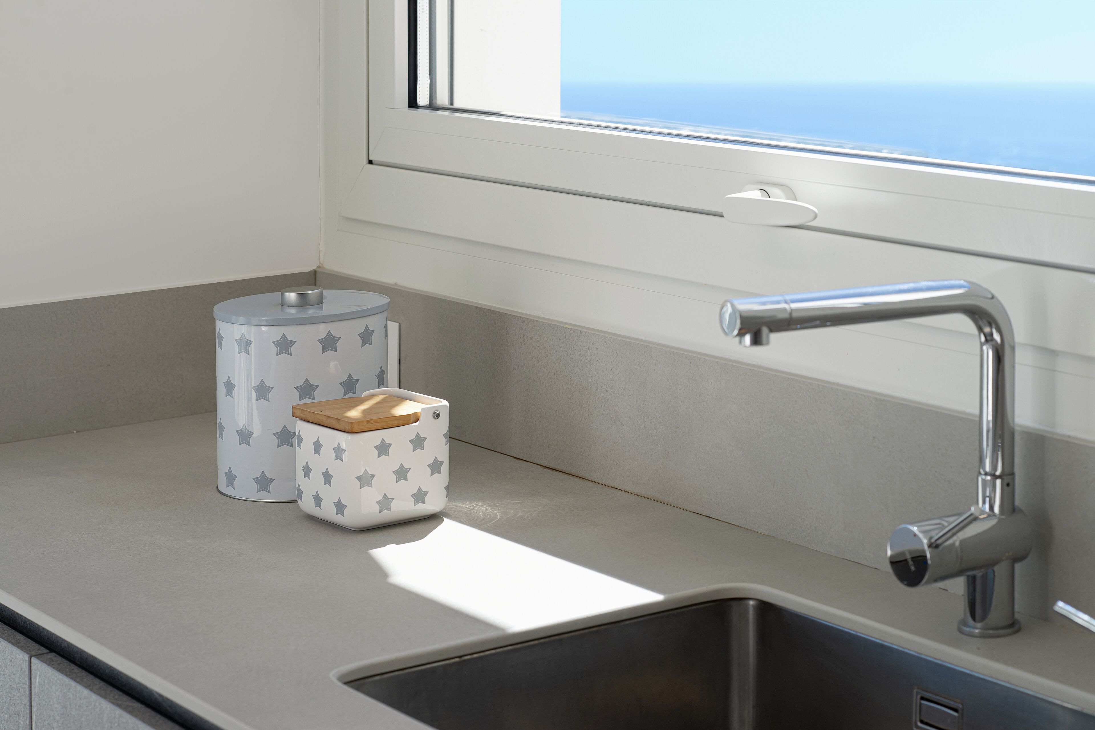 A kitchen with a stainless steel sink, a window with a view of the ocean, and a countertop with a star-patterned container and a wooden lid.