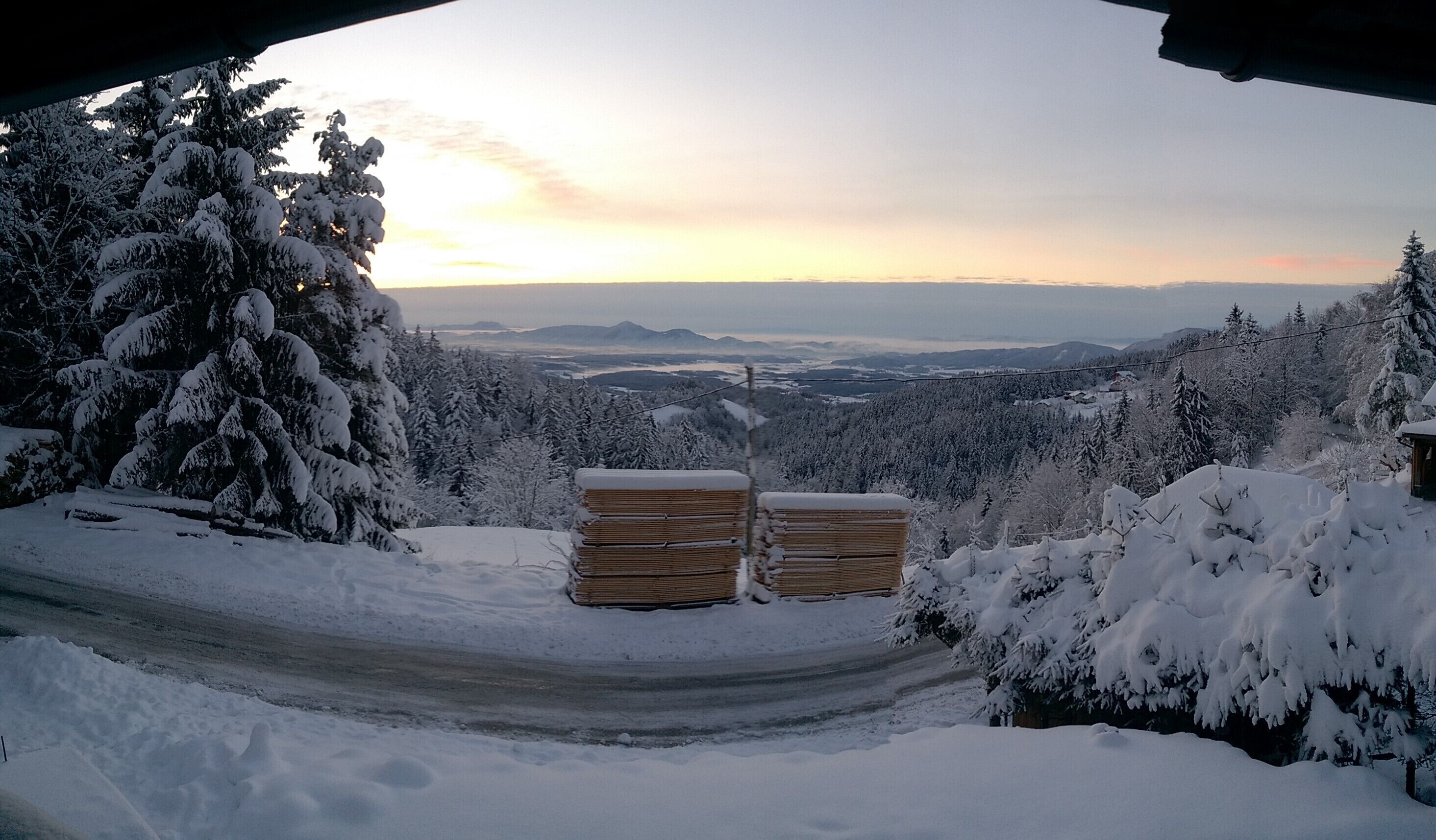 Schneebedeckte Landschaft mit einer Straße, Holzstapeln und verschneiten Bäumen.