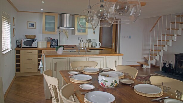 Un coin repas avec une table en bois dressée pour quatre personnes, entourée de chaises. On aperçoit, au fond, une cuisine équipée d’un four à micro-ondes et d’une cuisinière.