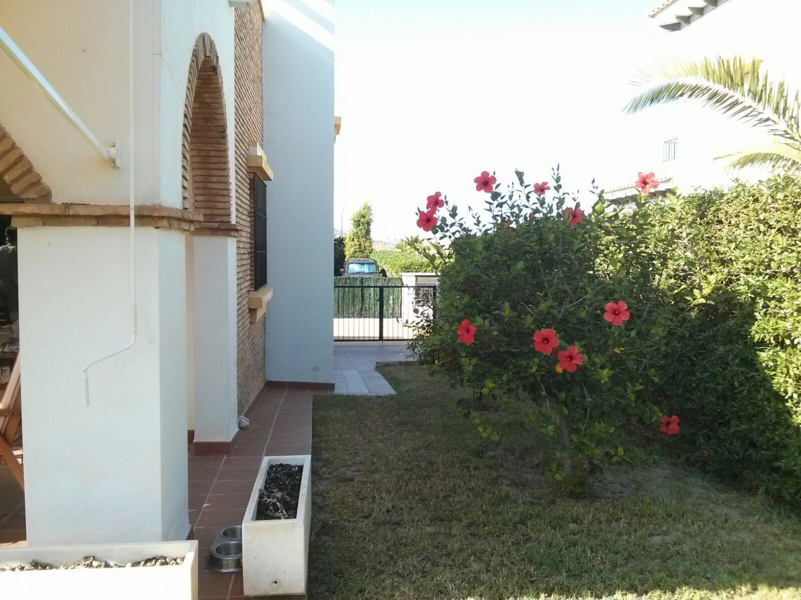 Ein Wohnhaus mit Ziegelbogen, einem kleinen Garten mit roten Blumen und einem Metallzaun im Hintergrund.