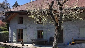Une maison en pierre avec un toit rouge, un banc en bois à l’extérieur et un arbre devant.