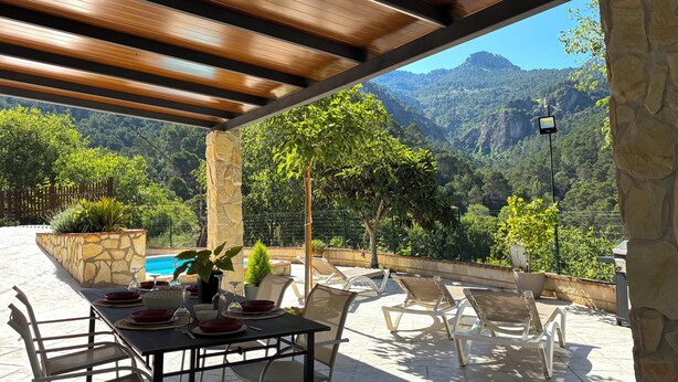 A covered outdoor dining area with stone walls, a pool, and a mountainous backdrop.