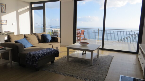 A modern living room with a sofa, coffee table, and large windows offering a view of the sea. - Apartment Goncalves,  (Arco da Calheta)
