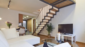 A modern living room with a white sofa, a glass coffee table, a TV on a white stand, a staircase, and a potted plant.