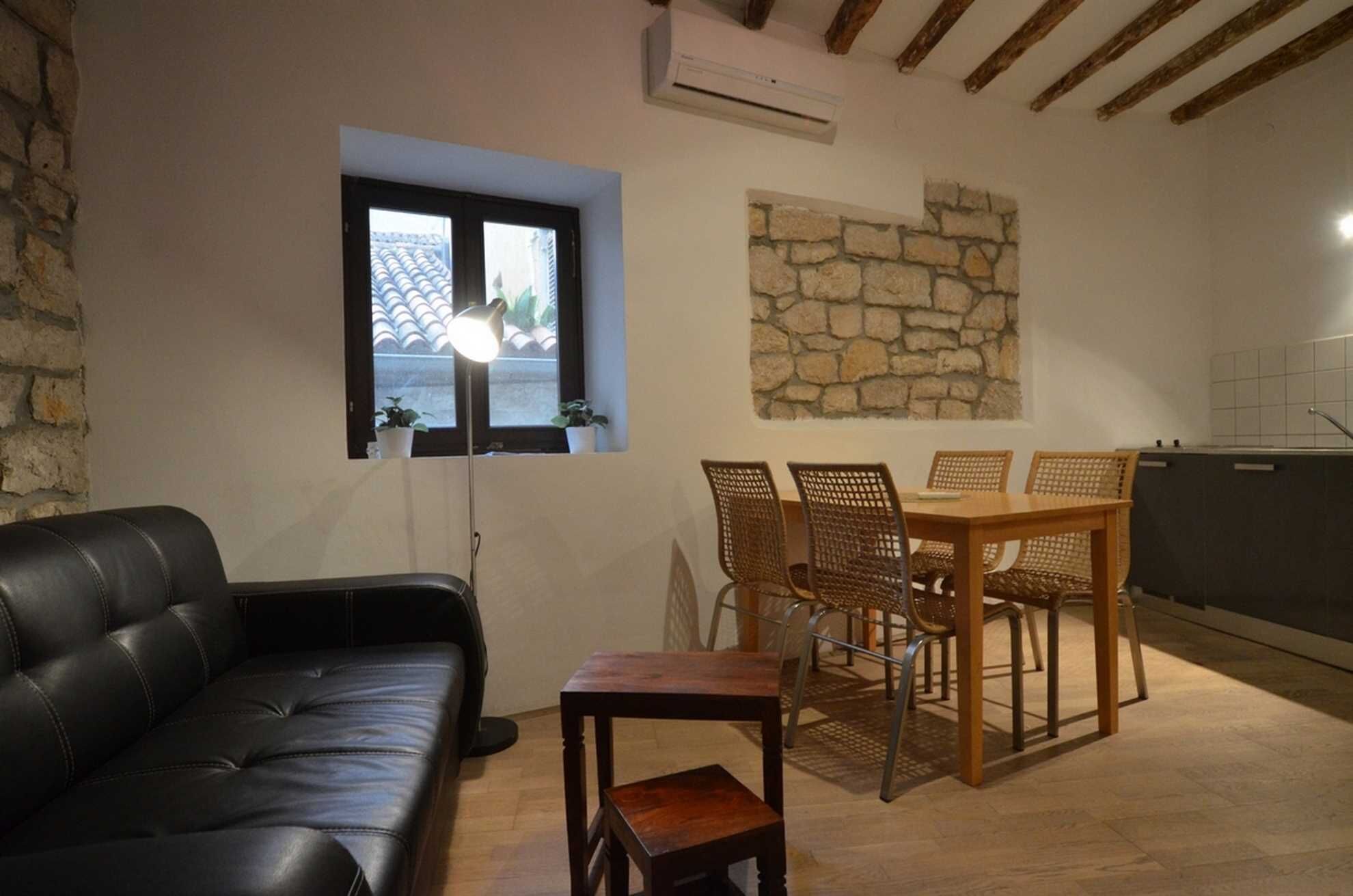 A cozy living room with a black leather sofa, a dining table with wicker chairs, a stone wall, and a window with a view of a tiled roof.