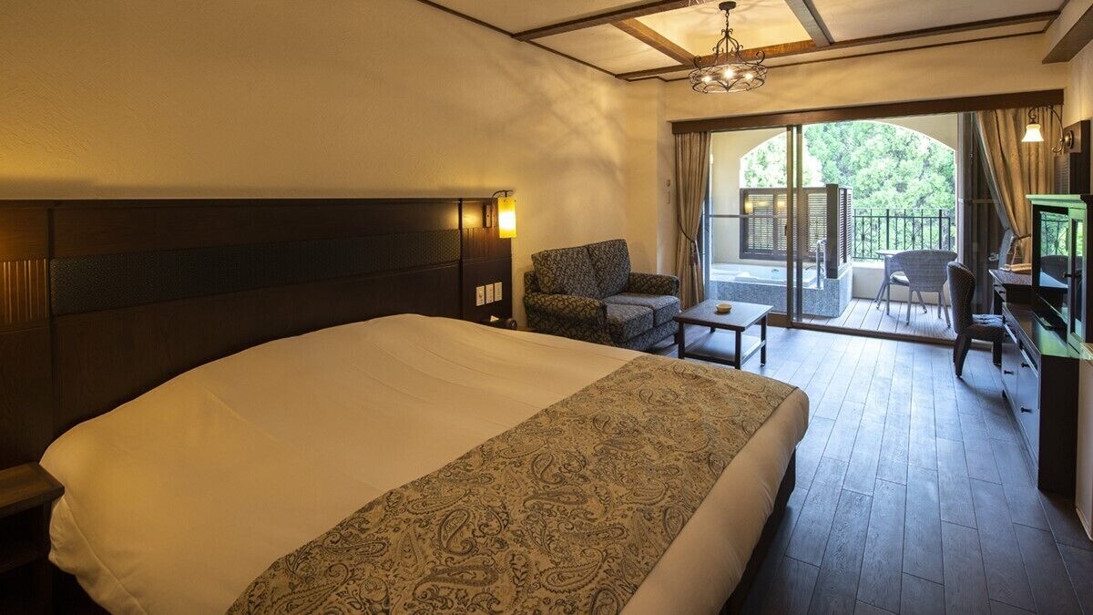 A hotel room with a large bed, a chair, a small table, a TV, and a balcony with a view of greenery.