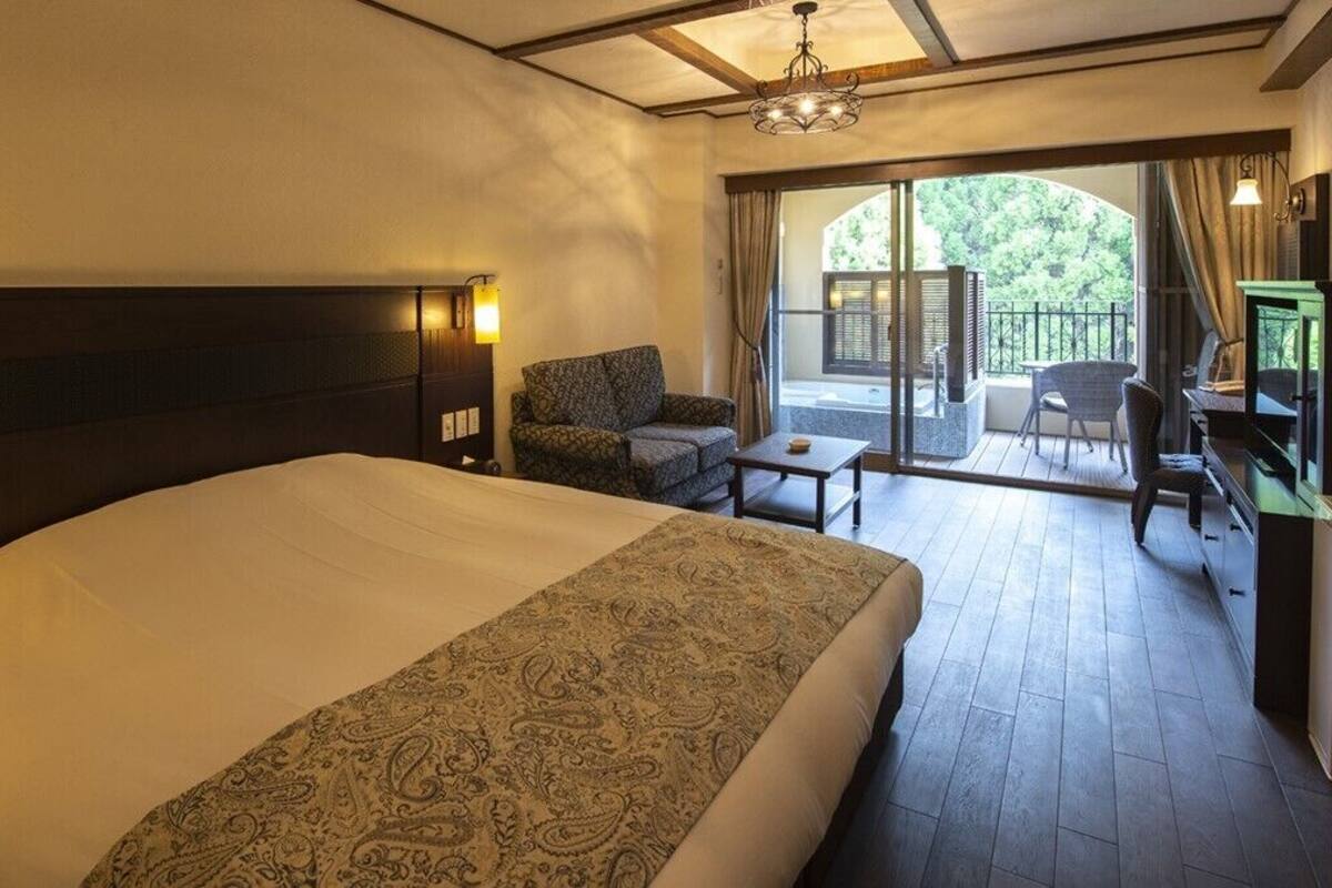 Hotelkamer met een groot bed, een stoel, een tafeltje, een televisie en een balkon met uitzicht op groen.