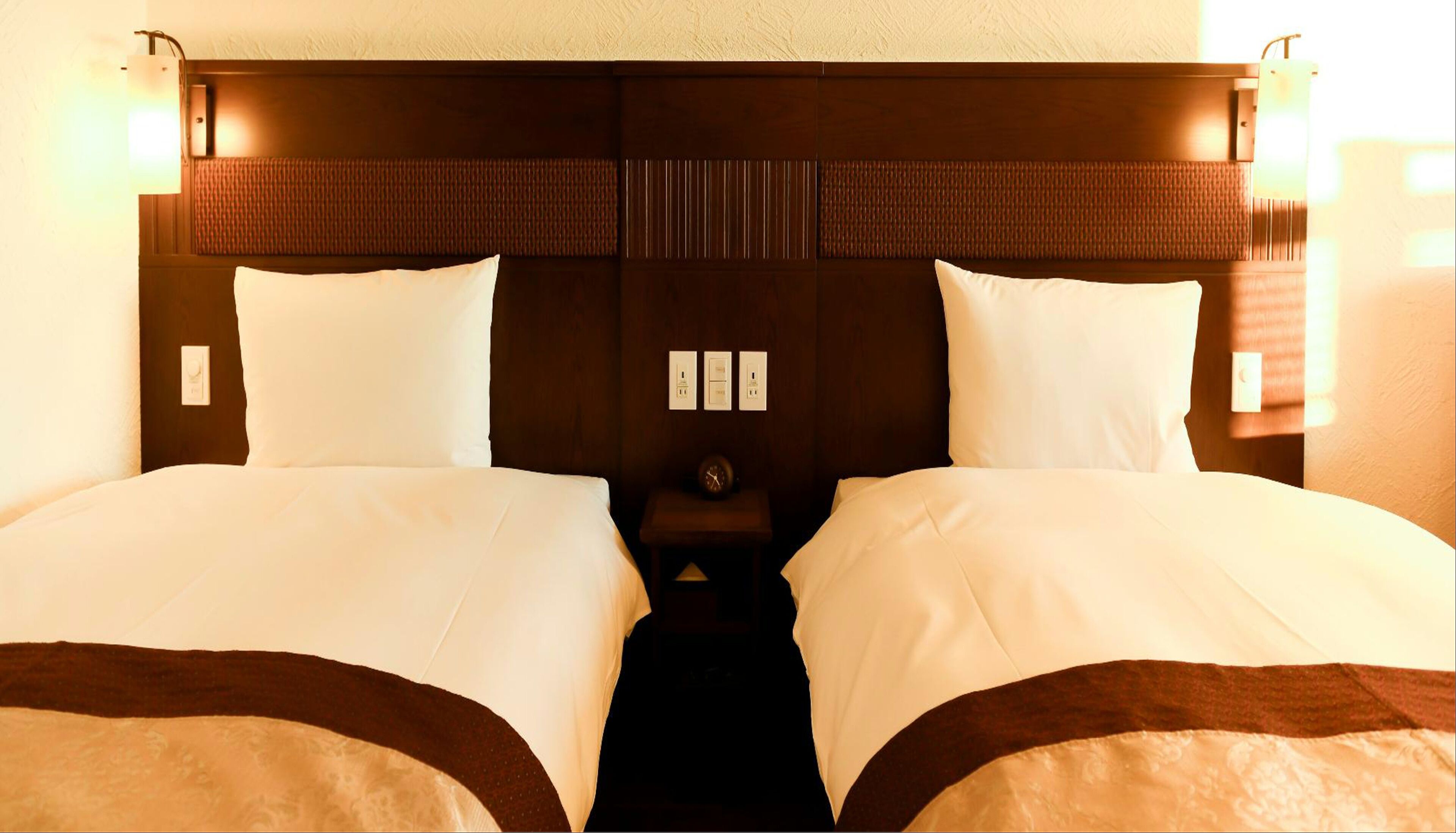 A hotel room with two beds, a nightstand with a clock, and wall-mounted light fixtures.
