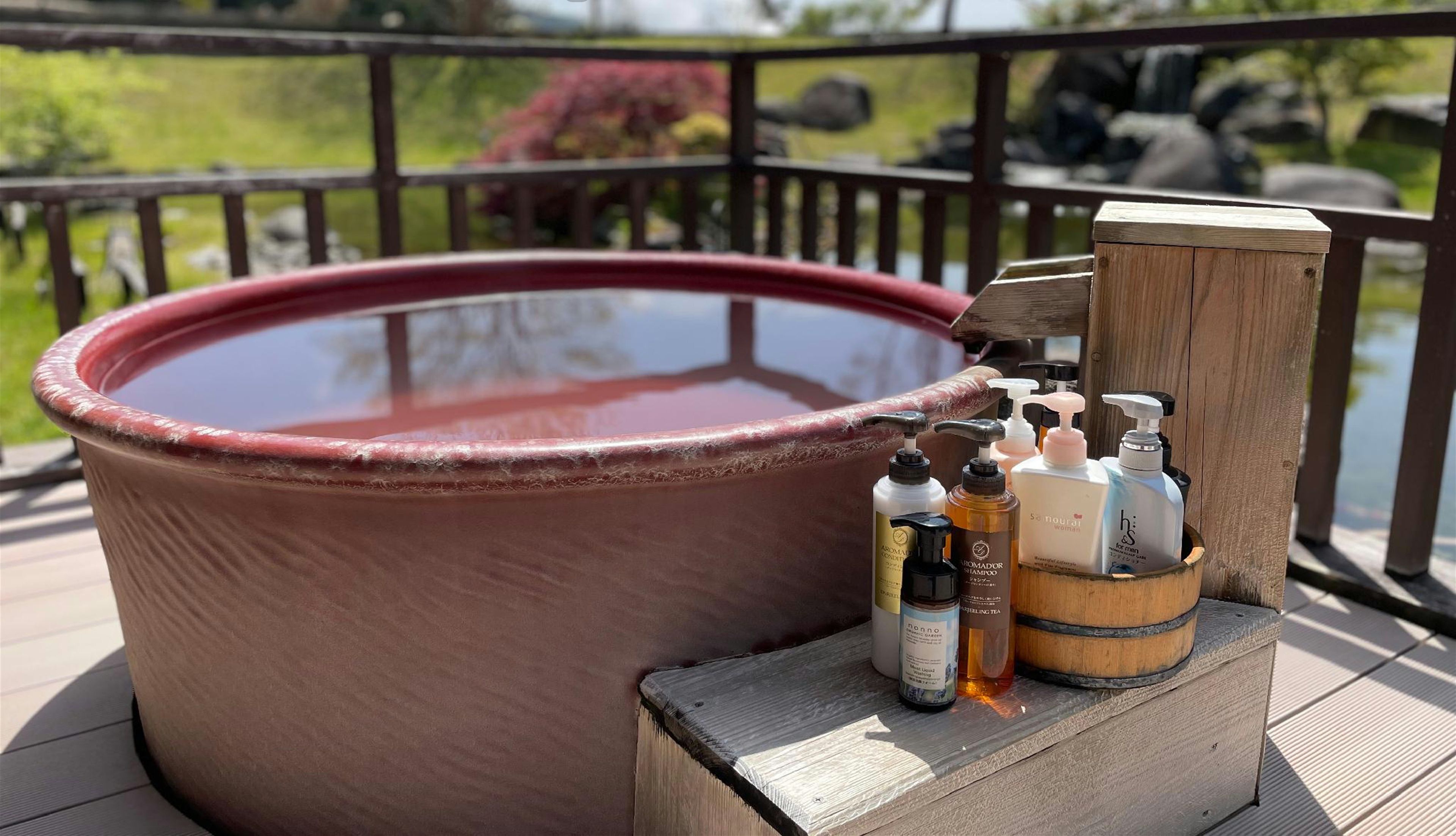An open-air bath with a wooden deck and a view of greenery in the background.